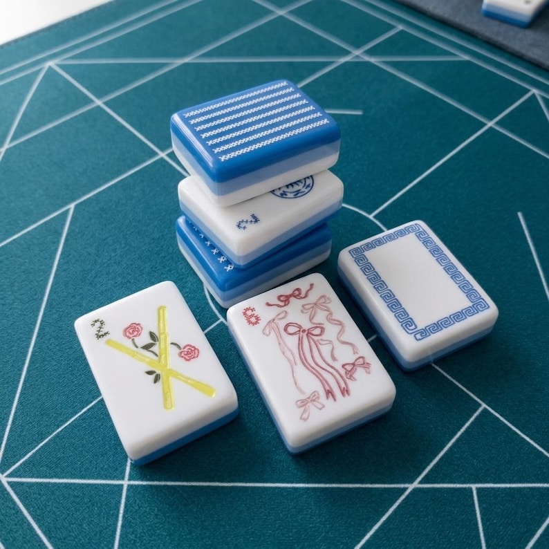 Op de afbeelding: Een verzameling rechthoekige mahjongtegels met witte voorzijden en blauwe achterzijden. E&eacute;n tegel heeft een ontwerp van gele bamboe en rode rozen met het cijfer 2, een andere toont roze linten en &eacute;&eacute;n heeft een blauwe geometrische rand. Drie tegels zijn opgestapeld.