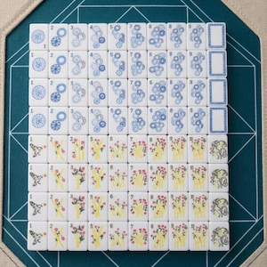 Op de afbeelding: Een set witte rechthoekige mahjongtegels gerangschikt in een raster. De bovenste vier rijen bevatten blauwe cirkelvormige en tandwielachtige patronen met de cijfers &eacute;&eacute;n tot en met negen en een lege tegel. De onderste rijen tonen kleurrijke bloem- en vogelillustraties.