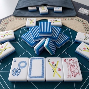 Op de afbeelding: Een set witte Mahjong-tegels met blauw-wit gestreepte achterkanten, voorzien van diverse kleurrijke illustraties waaronder bloemen, linten en geometrische patronen. De tegels zijn gerangschikt op een donkergroene mat met witte lijnen.
