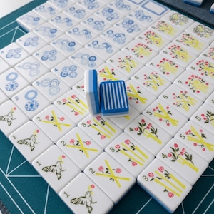 Op de afbeelding: Een set witte rechthoekige mahjongtegels met blauwe en gele prints, waaronder vogels, bloemmotieven en genummerde bamboestengels. Twee tegels staan rechtop in het midden, waarvan er &eacute;&eacute;n een blauw gestreept patroon op de achterkant toont.