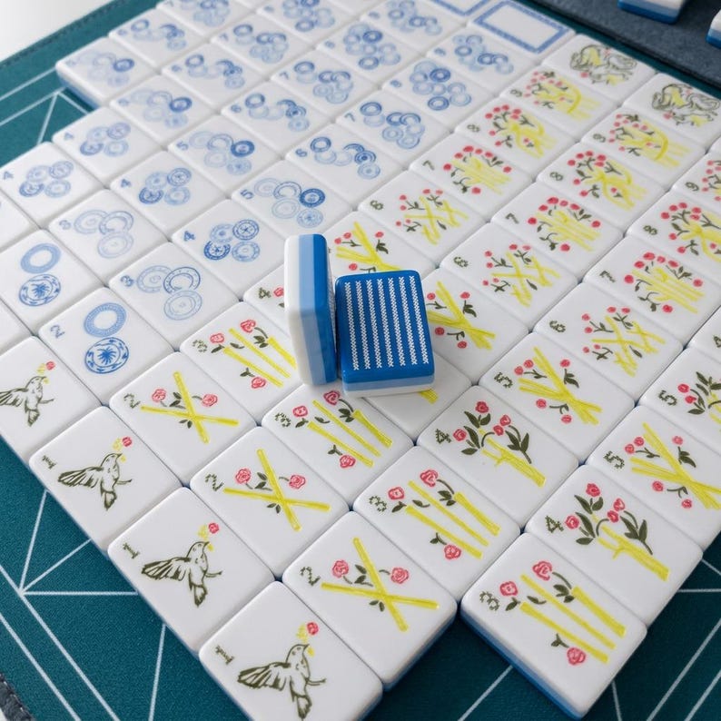 Op de afbeelding: Een set witte rechthoekige mahjongtegels met blauwe en gele prints, waaronder vogels, bloemmotieven en genummerde bamboestengels. Twee tegels staan rechtop in het midden, waarvan er &eacute;&eacute;n een blauw gestreept patroon op de achterkant toont.