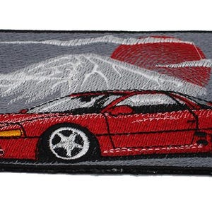 May include: A rectangular embroidered patch featuring a red sports car in profile against a gray background. Above the car, a white mountain peak is depicted with a large, solid red circular sun behind it, accented by thin white clouds.