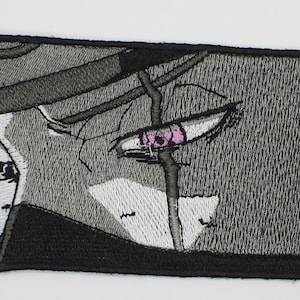 May include: A rectangular embroidered patch featuring a stylized black, white, and gray manga-style illustration of a face with a prominent eye. The eye is detailed with a small pink iris, and the design is framed by a thick black stitched border.