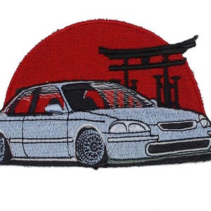 May include: An embroidered patch featuring a light blue hatchback car parked in front of a black torii gate silhouette against a solid red circular background. The car has detailed wheels and a low profile, with black stitching outlining the entire design.