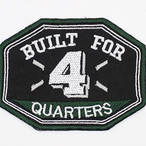 Op de afbeelding: Een achthoekige geborduurde patch met een zwart midden en een donkergroene rand. De tekst BUILT FOR is in het wit geborduurd boven een groot wit cijfer 4, met het woord QUARTERS in het wit eronder. Vier kleine witte streepjes omringen het cijfer 4.
