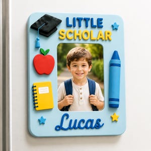 May include: A light blue rectangular photo frame featuring the text Little Scholar and Lucas. The frame is decorated with a black graduation cap, a red apple, a yellow spiral notebook, a blue crayon, and two small stars in blue and yellow.