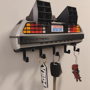 Puede incluir: Un soporte de pared para llaves con forma de parte trasera de coche deportivo, con rejillas negras, luces traseras rojas y naranjas, y una matrícula de California que dice OUTATIME. La unidad gris y negra incluye cinco ganchos negros.
