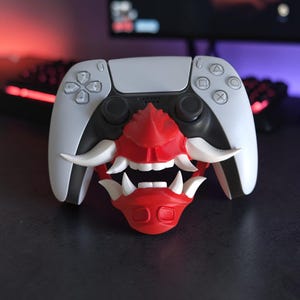Könnte beinhalten: Ein weißer Videospiel-Controller mit einem rot-weißen Maskenaufsatz, der die Mitte bedeckt. Die Maske zeigt ein stilisiertes Gesicht mit markanten weißen Fangzähnen, einem roten Nasenbereich und einem roten Kinnteil, das fest auf der Vorderseite sitzt.