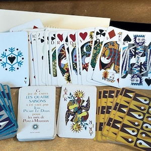 May include: A deck of playing cards titled Les Quatre Saisons, featuring custom illustrations on the face cards and backs. The set includes a white box, a joker card with a sun motif, and cards with blue and gold geometric patterns on the reverse.