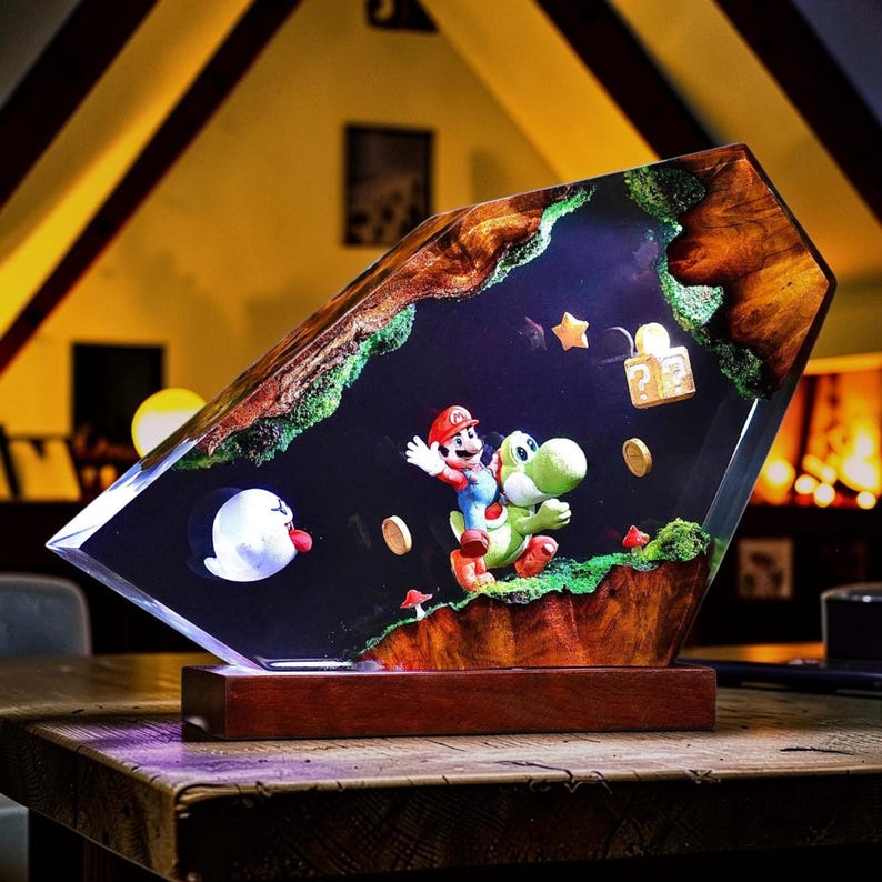 May include: A decorative resin lamp featuring a Mario and Yoshi figurine scene embedded within a clear, angular block with natural wood edges and green moss accents. The display includes a floating question block, coins, a star, and a ghost character.