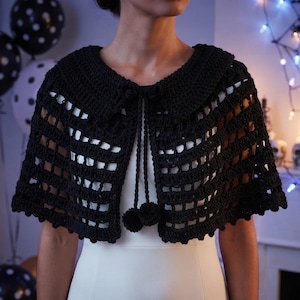 Peut inclure: Une capelette en crochet noir dotée d'un large col et d'une ouverture sur le devant. Le vêtement est réalisé avec un motif en grille répétitif et comprend deux longs cordons pendants qui se terminent par des pompons ronds au centre.