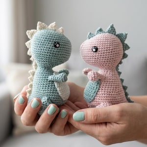 Peut inclure: Deux jouets dinosaures crochetés, un bleu et un rose, sont tenus dans des mains en coupe. Les dinosaures ont des dos épineux, de petits bras et de grands yeux. Le dinosaure bleu a des accents crème, tandis que le rose a des accents bleu-vert.