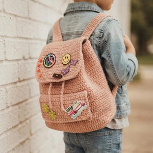 Peut inclure: Un sac à dos en crochet rose poudré doté d'un rabat orné d'un écusson signe de paix, d'un visage souriant jaune et de deux papillons violets. La poche avant est décorée de deux écussons fleurs vertes et d'un motif de flèches colorées.