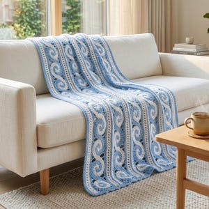 May include: A crochet blanket featuring a repeating pattern of light blue and white interlocking swirls and vertical stripes. The textile is draped over a cream-colored sofa, showcasing a textured, handmade design with consistent color blocks.