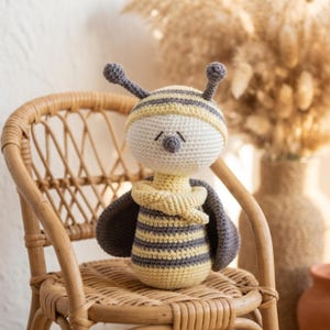 Peut inclure: Un jouet abeille en crochet avec une tête ronde blanche, un bonnet rayé jaune et gris, deux antennes et un corps cylindrique aux rayures horizontales jaunes et grises. L'abeille possède deux ailes grises et une petite écharpe jaune au cou.