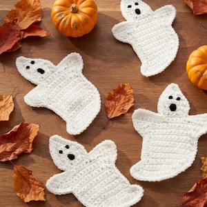 Peut inclure: Trois décorations fantômes en crochet blanc avec des yeux et des bouches en fil noir, disposées avec deux petites citrouilles orange et plusieurs feuilles d'automne séchées éparpillées. Les fantômes présentent un motif texturé plat.