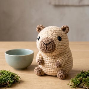Peut inclure: Un petit capybara en peluche crocheté de couleur beige, doté d'un museau, d'oreilles et de pattes marron foncé. La figurine possède des yeux ronds en plastique noir ainsi qu'un nez et une bouche brodés. Il est assis, avec une texture en laine.