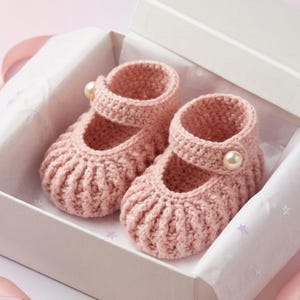 Peut inclure: Une paire de chaussons pour bébé en crochet rose clair avec une texture côtelée et une bride sur le dessus. Chaque chausson est orné d'un petit bouton rond en fausse perle de couleur crème sur le côté de la bride, présentés dans une boîte blanche.
