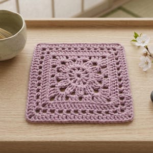 Peut inclure: Un dessous de verre carré au crochet de couleur mauve poussiéreux, présentant un motif floral central entouré d'une bordure géométrique. La pièce est réalisée avec un fil épais, mettant en valeur un design texturé avec des mailles visibles.