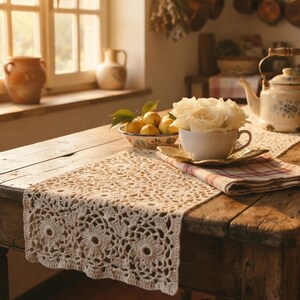 Peut inclure: Un chemin de table en crochet de couleur crème avec un motif floral répétitif est posé sur une table en bois. À côté se trouvent une tasse à thé blanche remplie de roses blanches sur une soucoupe, un bol de citrons jaunes et une théière blanche.