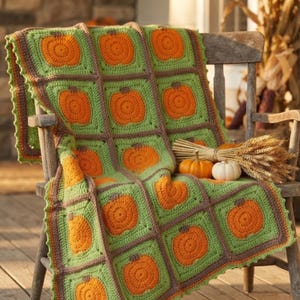 Peut inclure: Une couverture au crochet présentant un motif répétitif de citrouilles orange centrées sur des carrés vert clair, assemblés par des bordures en fil marron foncé. La couverture a un bord festonné et est posée sur une chaise en bois.