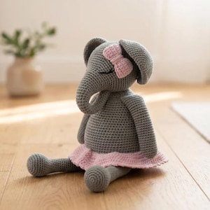 Peut inclure: Une poupée éléphant grise au crochet avec un nœud et une jupe roses. L'éléphant est assis sur un sol en bois, les jambes tendues. La poupée a les yeux fermés et une trompe. La jupe est de couleur rose clair.