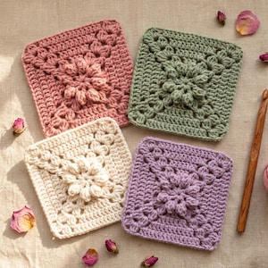 Peut inclure: Quatre motifs carrés au crochet en vieux rose, vert sauge, crème et lavande, chacun présentant un motif central de fleurs en points bouffants. Un crochet en bois est posé à droite, accompagné de pétales de rose séchés éparpillés.