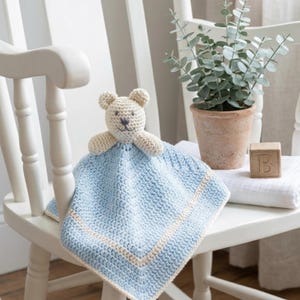 Peut inclure: Une couverture de sécurité au crochet avec une tête d'ours beige clair aux traits brodés, fixée sur une couverture carrée bleu clair bordée d'une bande crème. L'ours possède de petites oreilles arrondies et un museau simplement brodé.
