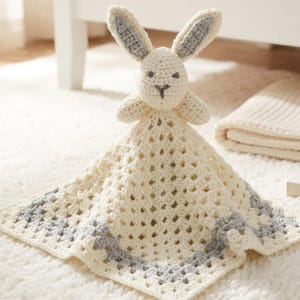 Peut inclure: Une couverture de sécurité au crochet avec une tête de lapin couleur crème aux longues oreilles et des accents gris à l'intérieur. La couverture attachée est en fil crème avec un motif carré et une bordure grise sur les bords extérieurs.