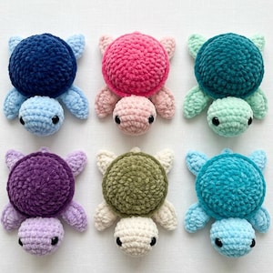 Op de afbeelding: Zes gehaakte schildpadknuffels gerangschikt in twee rijen. Elke schildpad heeft een getextureerd, rond schild en een kleinere kop met twee zwarte kraaloogjes. De kleuren zijn donkerblauw, roze, taling, paars, olijfgroen en helderblauw.