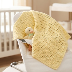 Peut inclure: Une couverture pour bébé en crochet jaune pâle avec un motif de points ajourés répétitifs, drapée sur un transat pour bébé blanc. Un petit canard en peluche avec un bec orange et un ruban vert clair autour du cou est glissé sous la couverture.