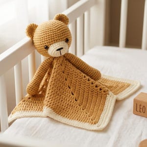Può includere: Una copertina di sicurezza all'uncinetto con una testa di orso color marrone dorato, occhi scuri e un muso color crema. La copertina attaccata è marrone dorato con un bordo color crema, caratterizzata da un motivo a punti e un design diagonale.