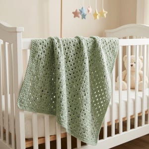 Peut inclure: Une couverture pour bébé en crochet vert sauge avec un motif de points granny square classique, drapée sur le côté d'un lit à barreaux blanc. La couverture présente un design texturé et ajouré avec un fil de poids constant et une teinte douce.