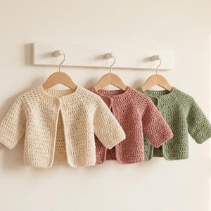 Peut inclure: Trois cardigans à manches longues en fil crocheté, suspendus à des cintres en bois. Les cardigans sont de couleurs unies : crème, vieux rose et vert sauge. Chaque vêtement présente un design simple ouvert sur le devant avec une encolure ronde.