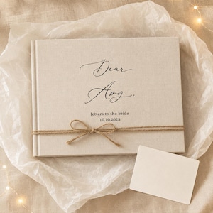 May include: A rectangular, light beige fabric-covered book with the black printed text "Dear Amy," "letters to the bride," and "10.10.2025." A thin, brown twine string is tied horizontally around the center of the book cover in a small bow.
