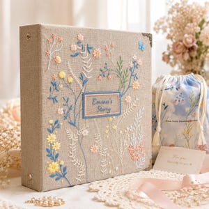 May include: A beige linen-covered photo album featuring intricate floral embroidery in blue, yellow, and pink threads. A central rectangular patch displays the embroidered text "Emma's Story" in blue, with metal corner protectors and a spine ring.