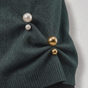Può includere: Un maglione verde scuro caratterizzato da un sottile motivo a maglia a spina di pesce. Il tessuto è arricciato sul lato e decorato con due perle sferiche lisce color crema e due perle sferiche dorate lucide e riflettenti di varie dimensioni.