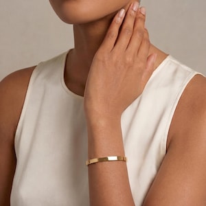 Puede incluir: Una pulsera rígida de tono dorado pulido con una superficie lisa y plana y un diseño de banda simple y continuo. La pulsera presenta una estética limpia con un acabado metálico reflectante que envuelve la muñeca en forma circular.
