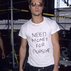May include: A white short-sleeved t-shirt featuring the black hand-written text NEED MONEY FOR PORSCHE on the chest. The shirt has a crew neckline and a slightly distressed collar. The garment is paired with dark trousers and a silver-toned watch.