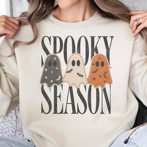 May include: A sand-colored crewneck sweatshirt featuring a graphic print of three ghosts in black, cream, and orange. The ghosts are decorated with stars, moons, and bats. The text "SPOOKY SEASON" is printed in a bold, black, distressed serif font.
