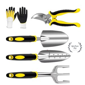 May include: A gardening tool set featuring a pair of black and yellow gloves, pruning shears, two trowels, and a hand rake. One trowel has a printed scale showing 10 cm and 4 inches. The set includes black and yellow handles on all metal tools.