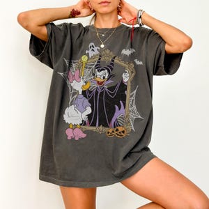 May include: A charcoal gray short-sleeve t-shirt featuring a graphic print of a duck character dressed as a villain inside an ornate gold frame. The design includes a white duck, a ghost, spiderwebs, bats, and two orange jack-o'-lanterns at the base.
