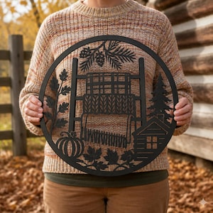 May include: A circular black wall hanging featuring a cutout design of a ladder with a draped blanket, a pumpkin, oak leaves, pine branches with cones, a pine tree, and a small cabin. The piece is crafted with intricate, flat, silhouette-style details.