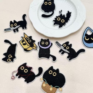 Peut inclure: Une collection d'écussons brodés représentant des chats noirs dans diverses postures, notamment avec des couteaux, dans des boîtes, emmêlés dans du fil rouge ou portant des colliers, avec de grands yeux jaunes et des détails noirs.