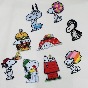 Peut inclure: Un ensemble de neuf écussons brodés représentant le personnage Snoopy dans diverses tenues et activités, notamment avec une perruque colorée, un casque de pilote, un bonnet de Noël, ou allongé sur un hamburger et dans une niche rouge.
