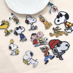 Peut inclure: Une collection d'écussons en tissu brodé représentant divers personnages Peanuts, dont Snoopy, Woodstock, Charlie Brown, Lucy et Dingo. Les écussons présentent des motifs colorés des personnages dans différentes poses et tenues sur un fond blanc.