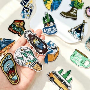 Peut inclure: Une collection d'écussons brodés sur le thème de la nature et du plein air, incluant une ampoule avec des montagnes, une lanterne, une chaussure de randonnée, un van, une tente et des tasses avec le texte Explore et Drink Coffee Outdoors.