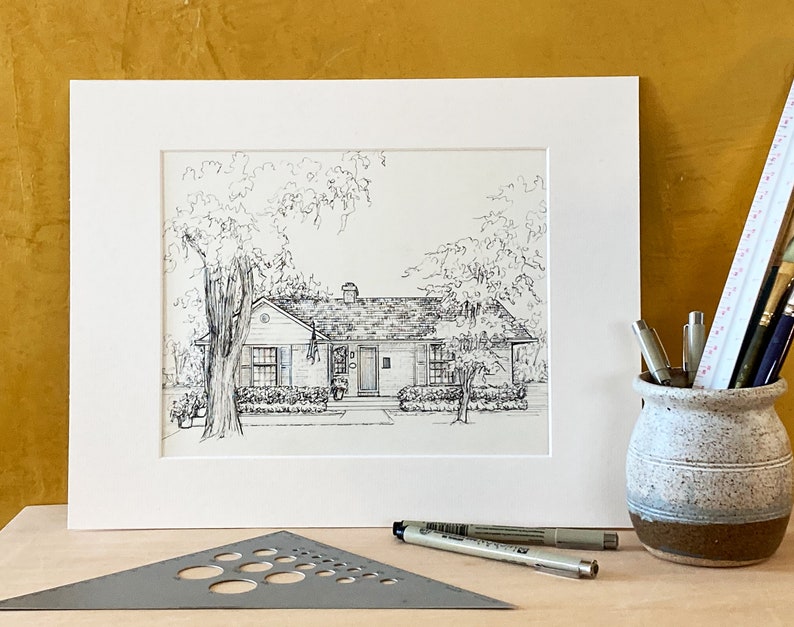 May include: A black and white line drawing of a house with a front yard and trees. The house has a gabled roof, a front porch, and a window on each side of the front door. The trees are drawn with a lot of detail, and the drawing is framed in a white mat.