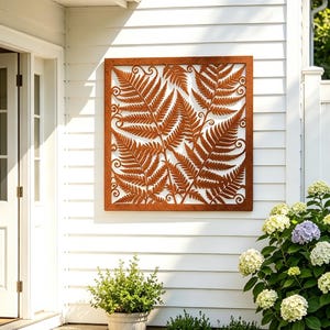 May include: A square metal wall art panel featuring a detailed laser-cut fern leaf pattern with a rusted brown finish. The decorative piece is mounted on a white horizontal siding wall, showcasing intricate foliage silhouettes and scrollwork.
