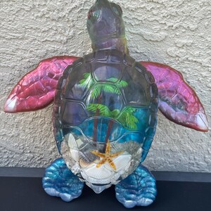May include: A translucent sea turtle figurine with iridescent pink flippers and blue legs. The shell contains a miniature diorama featuring green palm trees, sand, white seashells, and a small orange starfish encased in clear resin.